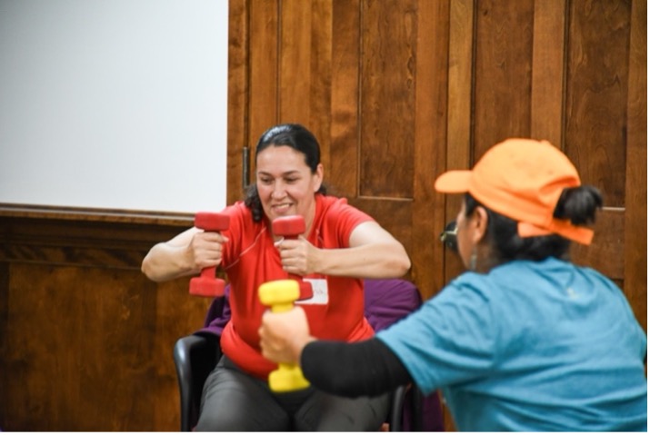 A seated GenteFuerte participant smiles as she brings her weights close together in front of her.