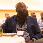 A father listens with other conference attendees to a keynote speaker during lunch.