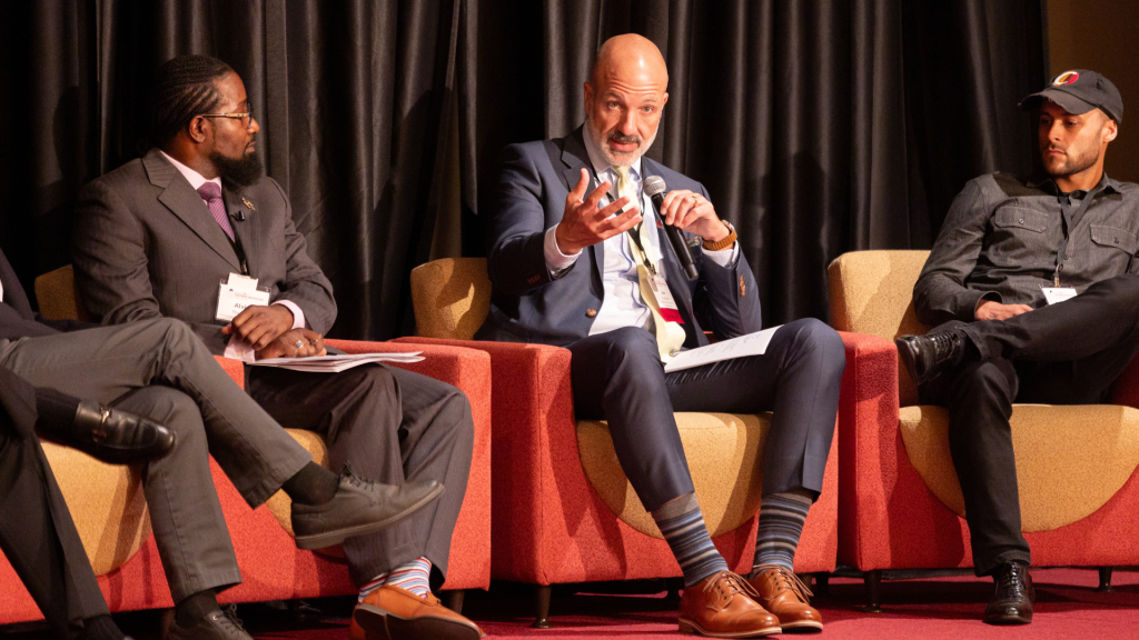 Three fathers sit in lounge chairs on a stage. The middle man is holding a microphone and talking to the audience.