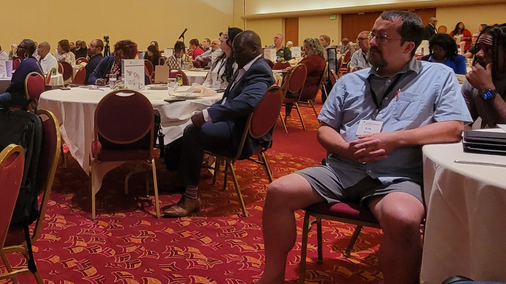 Participants at a conference sit at tables while listening to a speaker