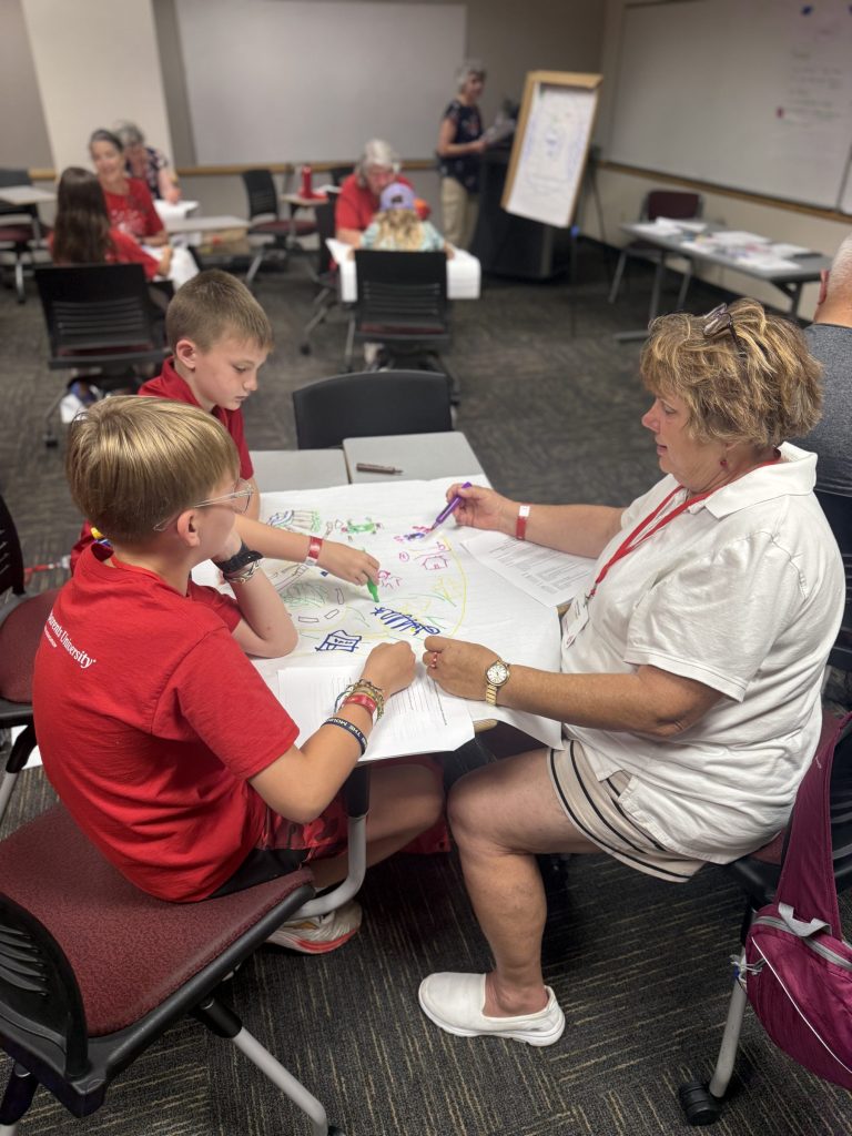 Woman and 2 boys sitting at a table drawing on a large piece of paper.