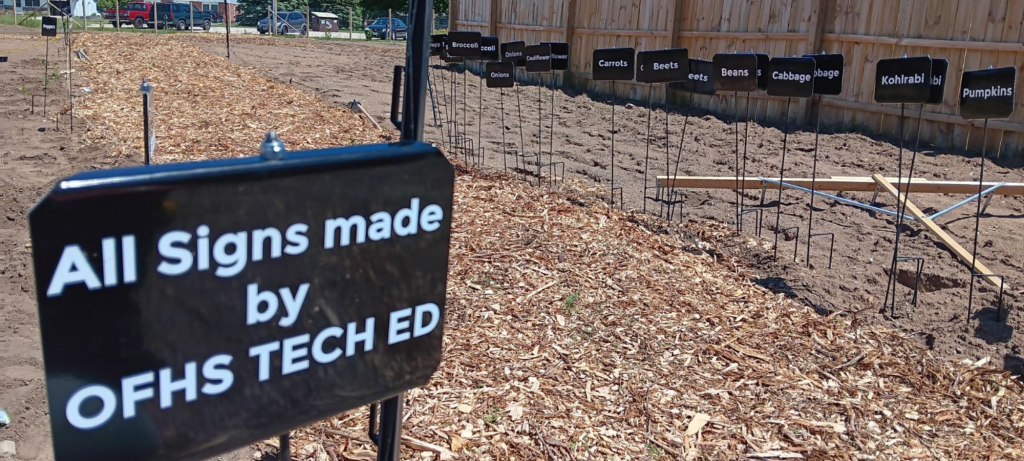 Black metal signs with vegetable names in white letters on wire stands in the ground, lined up in a patch of dirt. One sign says "All signs made by OFHS Tech Ed".