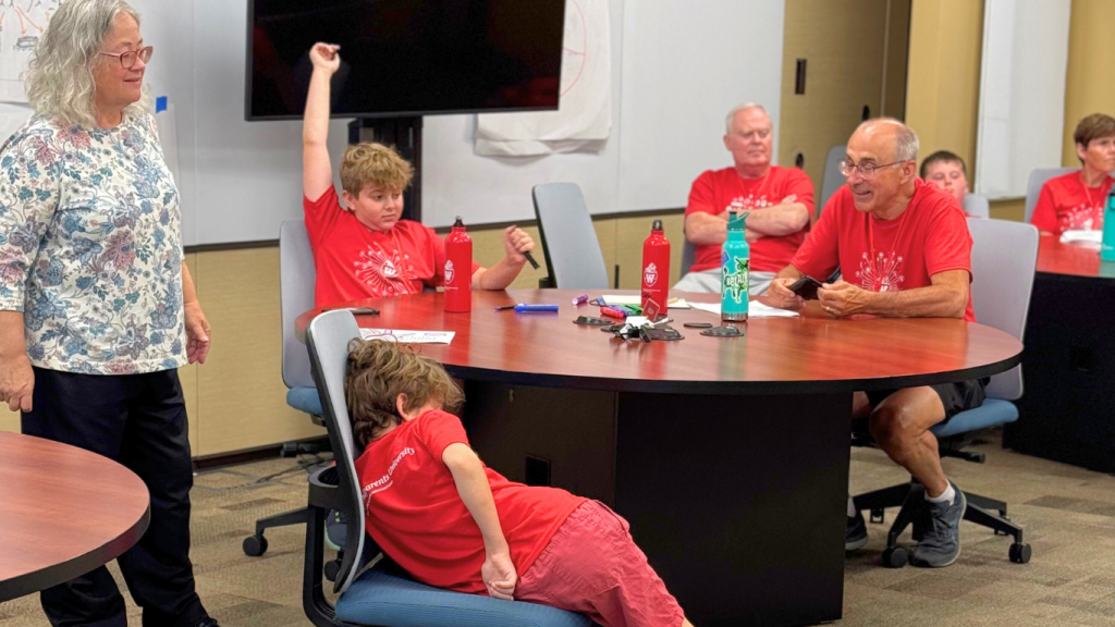 A grandfather and 2 boys sit at a table and discuss while the teacher stands beside them