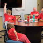A grandfather and 2 boys sit at a round table while the teacher stands beside them watching