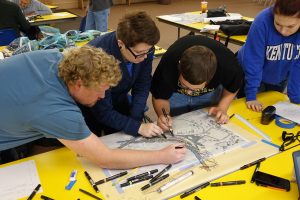 Three people stand over a large map on a table, drawing on the map with pens