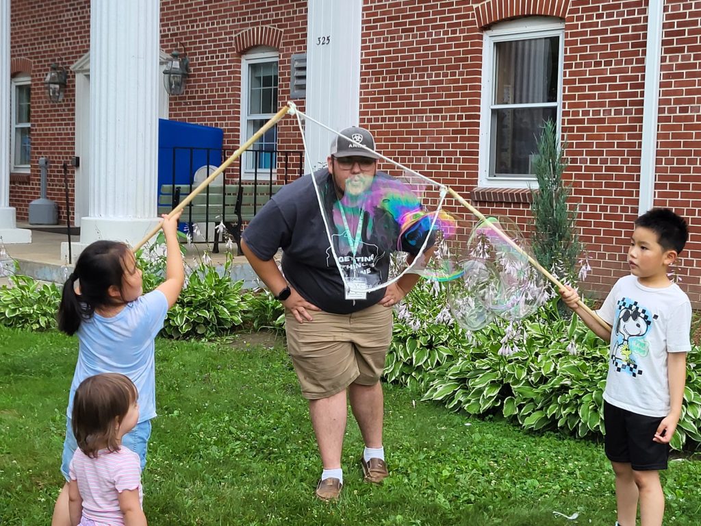 An AmeriCorps member blows a giant bubble in a big bubble wand held on each side by youth. They are on the lawn of the library for a bubble science program.