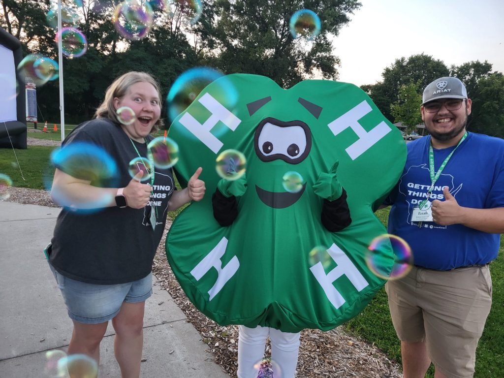 Two AmeriCorps members smile and give two thumbs up. They are standing on each side of a 4-H Clover mascot to show the partnership between AmeriCorp and 4-H.