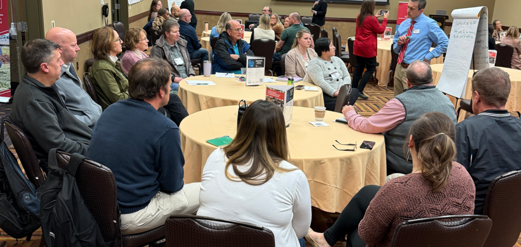Group of professionals sit around tables and listen to a woman talk.