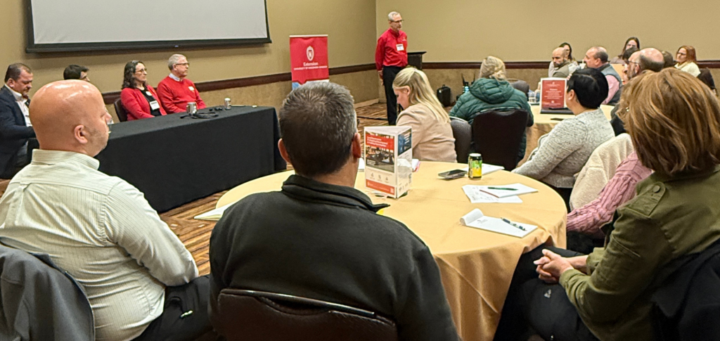 Group of people sit at a table in the front of a room, listening to an audience member talk.