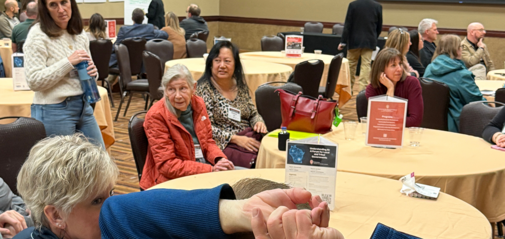 Group of people sit around circle tables listening to another person speak.