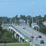 Bridge with cars driving over a river. Bridge is surrounded by green trees and interspersed with roofs of houses and a brick clock tower peeking over the tree line on the far side of the bridge.