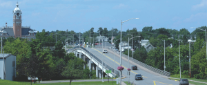 Bridge with cars driving over a river. Bridge is surrounded by green trees and interspersed with roofs of houses and a brick clock tower peeking over the tree line on the far side of the bridge.