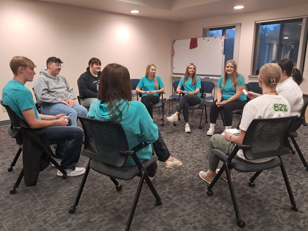 A group of nine young adults sit in a circle on rolling office chairs in a meeting room, engaging in a group discussion or activity. 