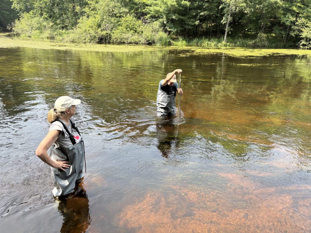 Two people stand in a river in waders. One person is in the middle of the river, pushing a long pole down to measure.