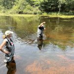 Two people stand in a river in waders. One person is in the middle of the river, pushing a long pole down to measure.