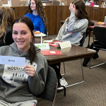 Two youth sit at tables in a council chamber smiling at the camera. The one on the left holds up a paper slip with the word "MOCK" printed on it. In the background, several other youth sit at tables with folders and name tags, interacting in a group discussion.