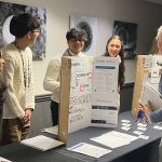 Students stand behind a table with a poster board explaining the poster's contents to a person standing in front of the table