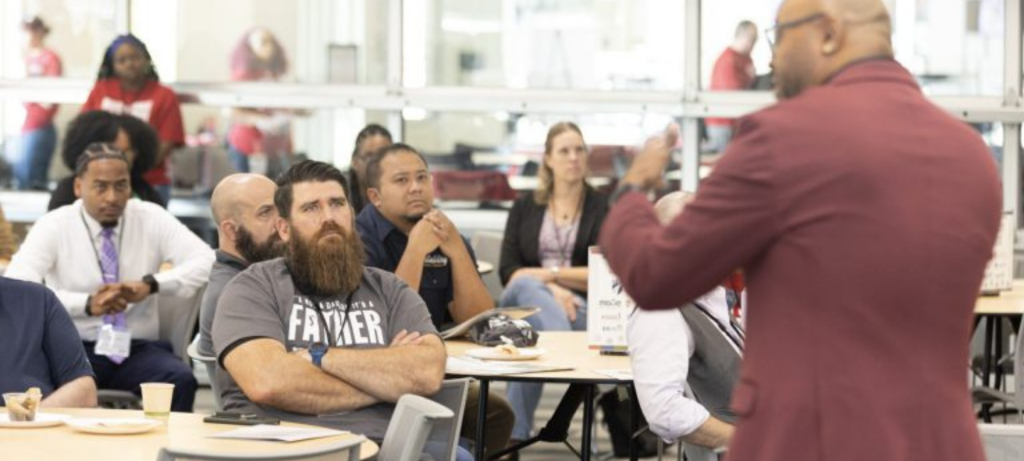 Classroom with a group of people sitting at tables, listening to the speaker. One person's tshirt says "Father."