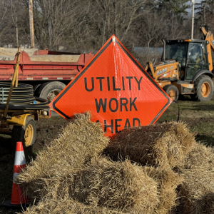 Orange diamond-shaped road sign surrounded by bales of hay that says "Utility Work Ahead"
