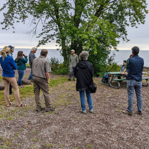 Group of adults stand outside listening to speaker near the shore of Lake Superior