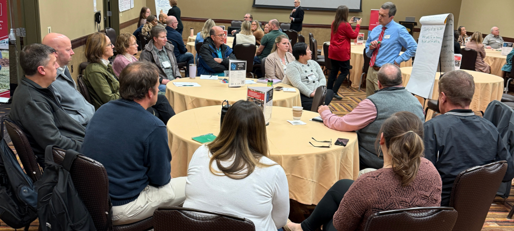 Groups of people sit around round tables facing a facilitator standing beside a large pad of paper