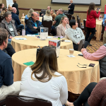 Groups of people sitting at round tables, looking at facilitators writing on a large pad of paper