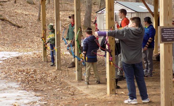 This image shows three children standing under a structure supported by wooden beams. All of the children are holding archery bows. The closet child has the string pulled back and is aiming the arrow.