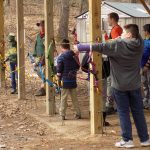 This image shows three children standing under a structure supported by wooden beams. All of the children are holding archery bows. The closet child has the string pulled back and is aiming the arrow.