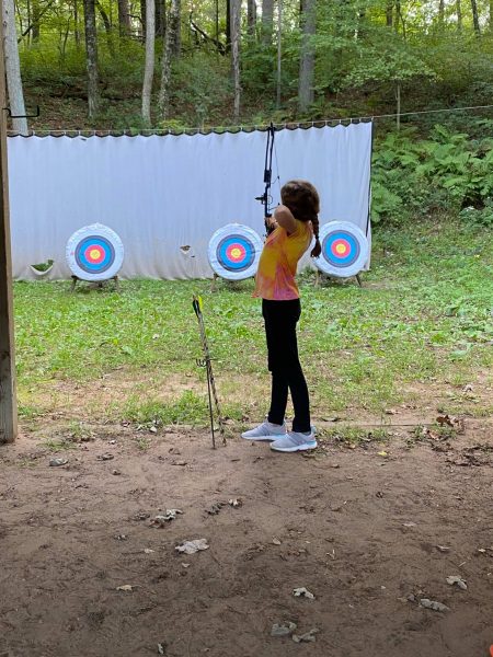 This image shows a young girl in a tie-dye shirt holding a bow in the foreground. There are 3 circular targets in the background.