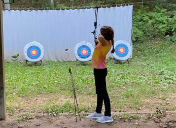 This image shows a young girl in a tie-dye shirt holding a bow in the foreground. There are 3 circular targets in the background.
