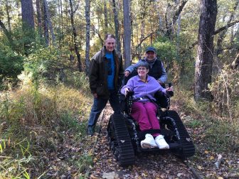 Three people are next to each other smiling at the camera. The person in the middle is sitting in an outdoor wheelchair.