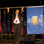 Image of George Koepp holding up the Wisconsin Flag at the NACAA AM-PIC flag ceremony. George is wearing a black suit and red, white & blue tie.